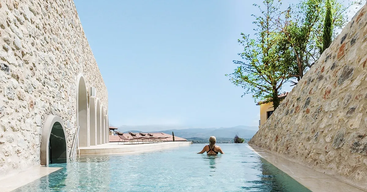 A blonde woman overlooks rolling countryside while standing in an outdoor pool on a sunny day at Euphoria Spa, Mystras, Greece.