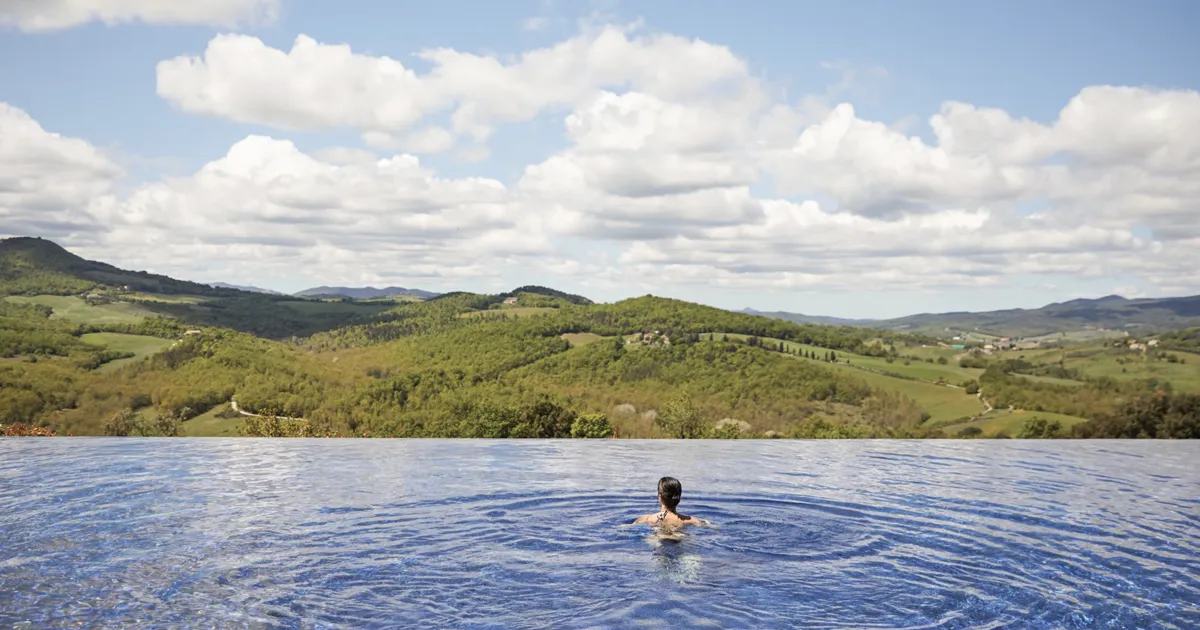 A brunette woman relaxes in an infinity pool overlooking forested green valleys in Tuscany, Italy.