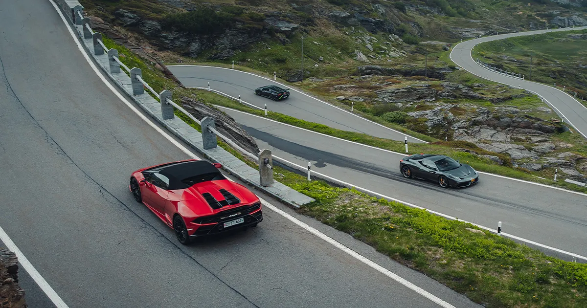 A red Lamborghini leads a grey Ferrari in ascending a steep pass on a supercar tour.
