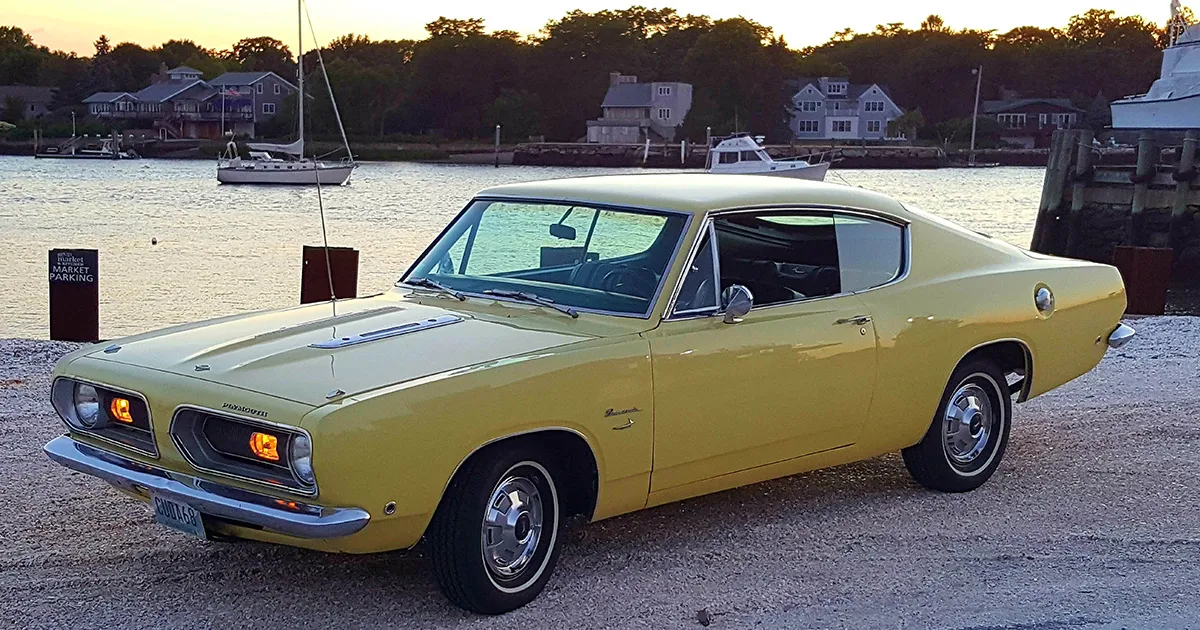 A Plymouth Barracuda in mustard paint next to the ocean at sunset.
