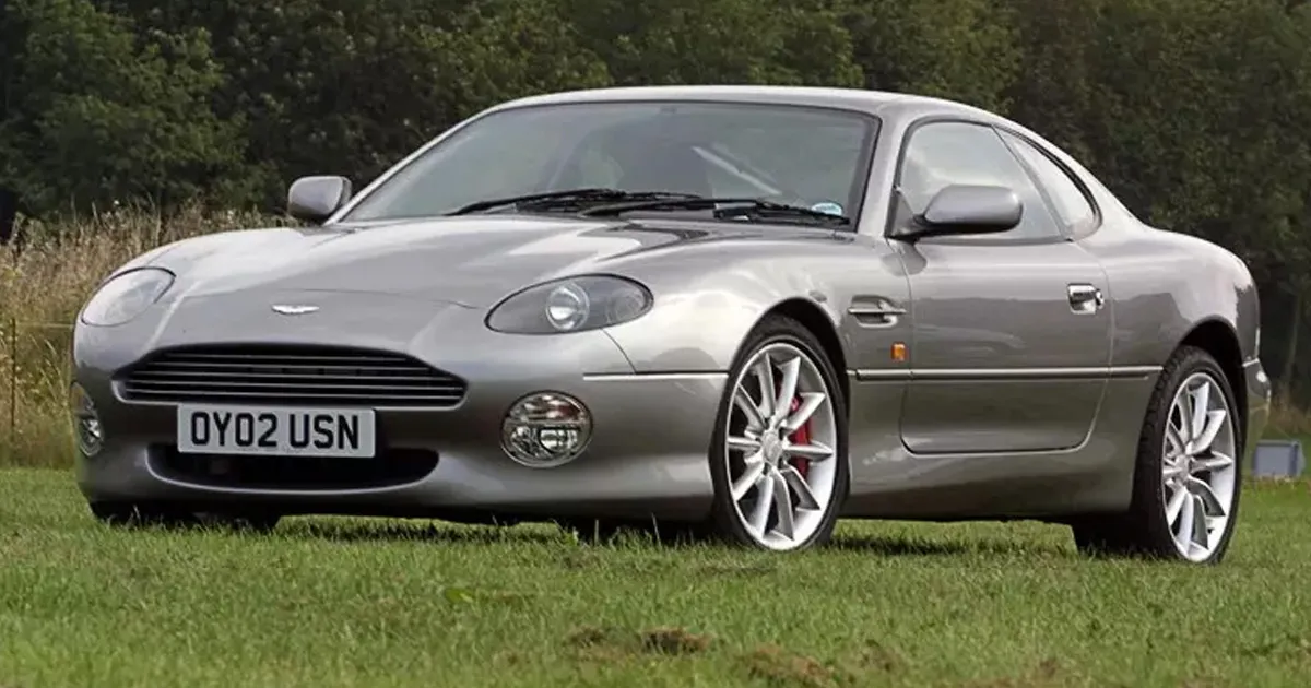 A dark silver Aston Martin DB7 coupe sitting on grass.