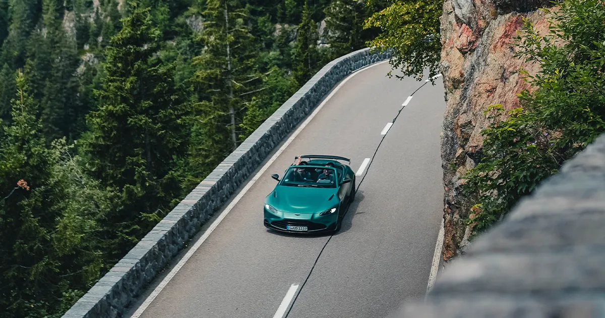 A teal Aston Martin Vantage F1 Roadster being driven with its top down by two Ultimate Driving Tours guests.