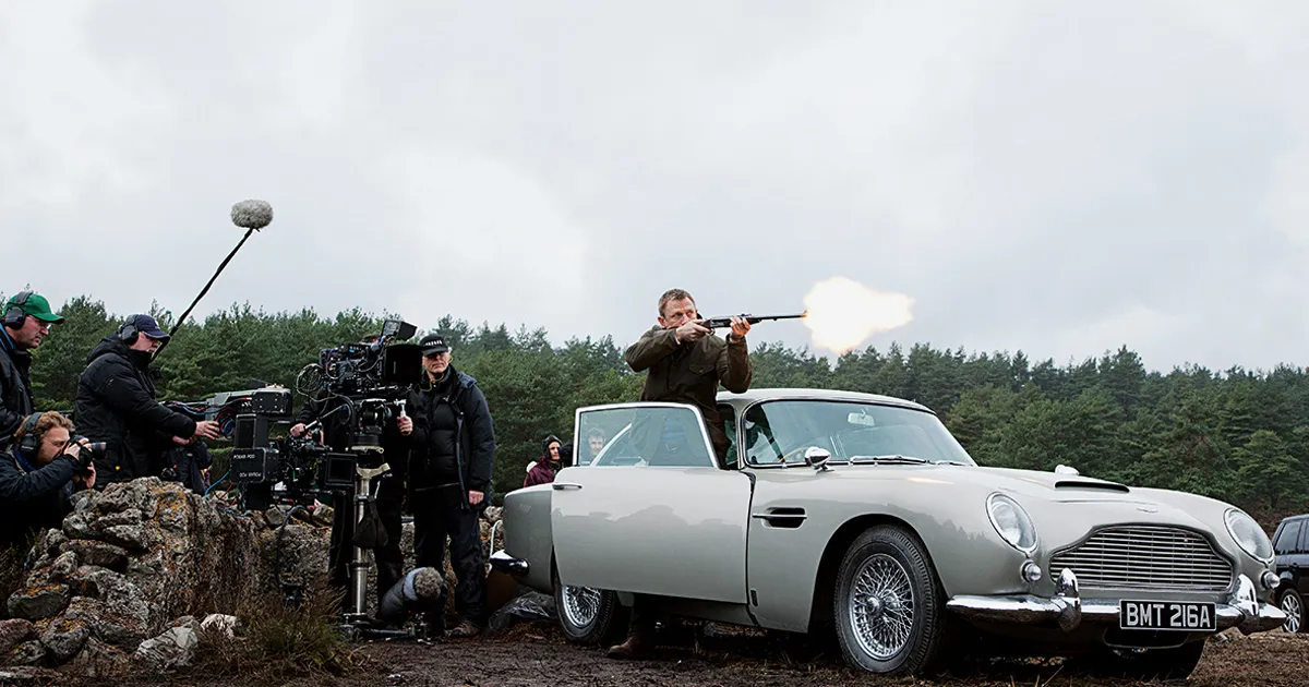 A film crew records Daniel Craig as James Bond as he fires a gun from behind an Aston Martin DB5.
