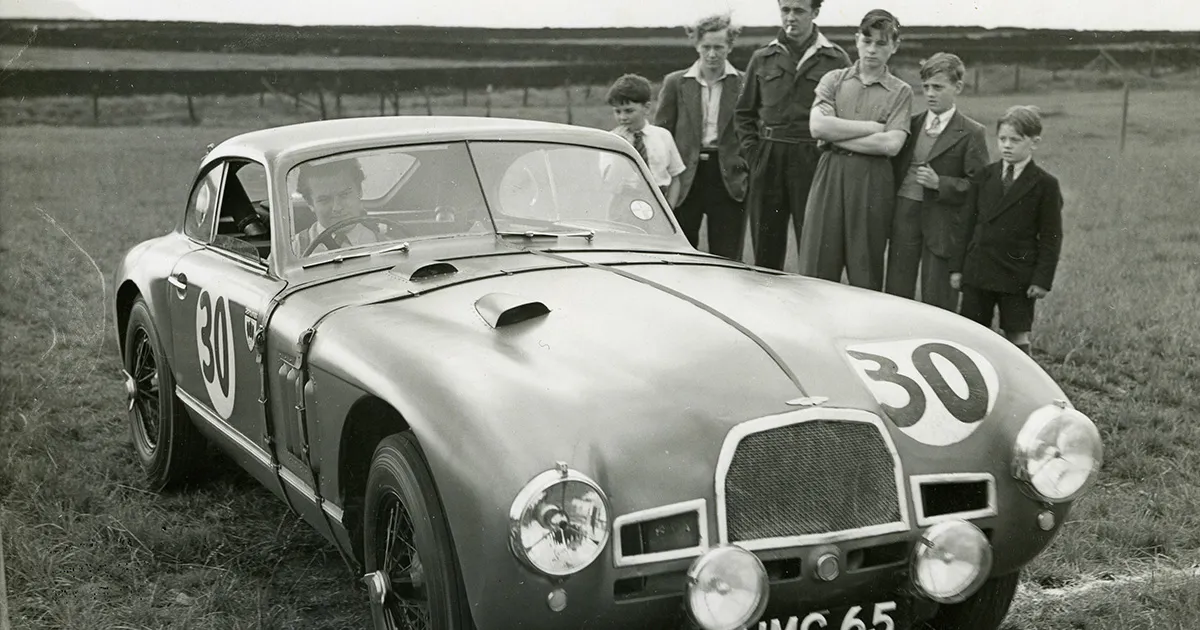 A man drives a vintage Aston Martin through a paddock as a small group in formal attire watches on.