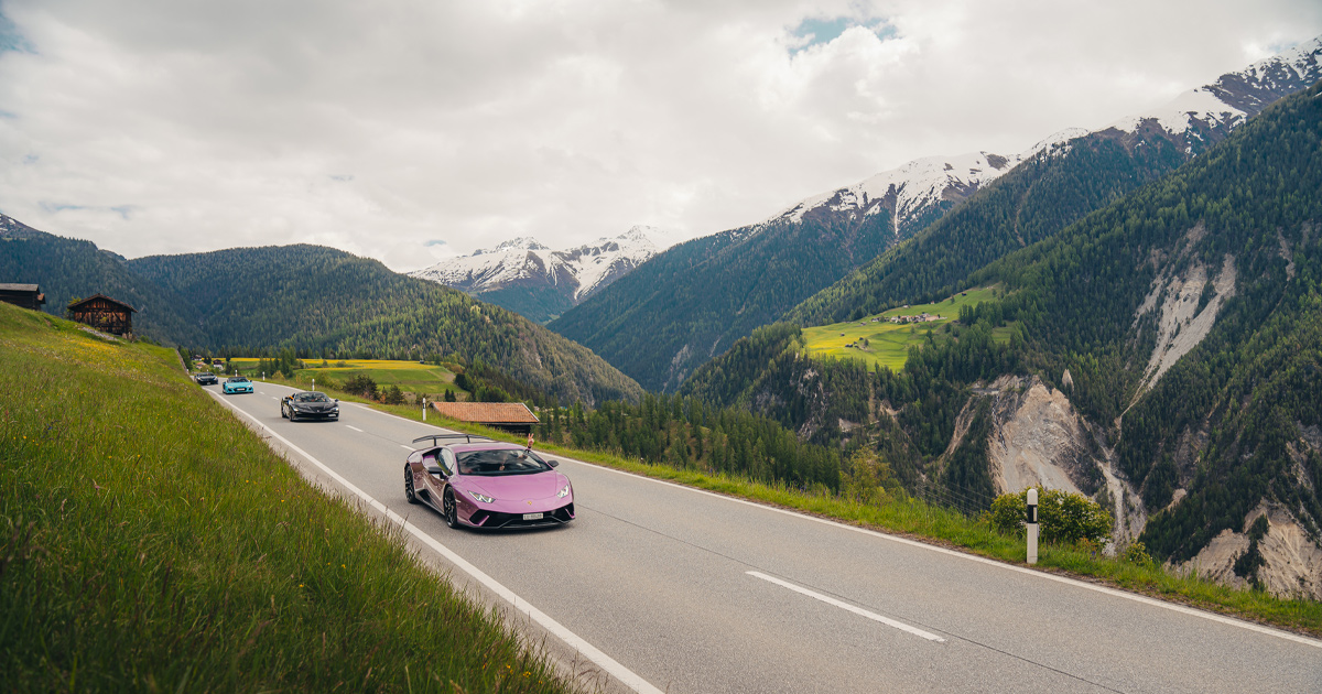 A purple Lamborghini Huracan leads a convoy of supercars along an alpine road in Europe
