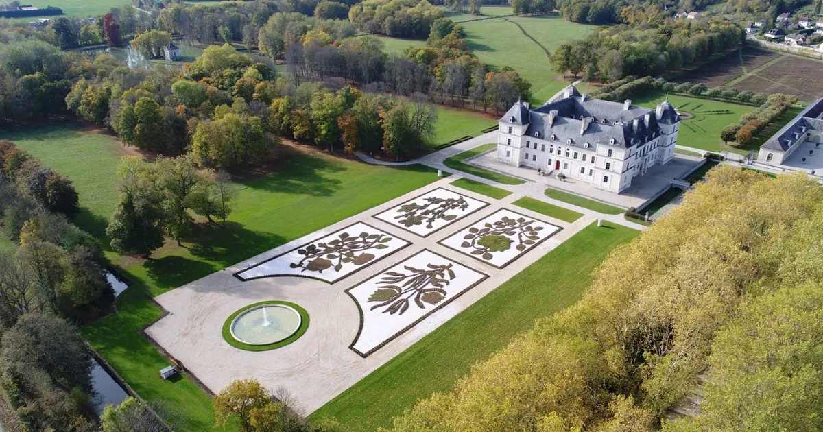 A huge white stone château in Burgundy with decorative flowerbeds and a large fountain within its sweeping grounds.