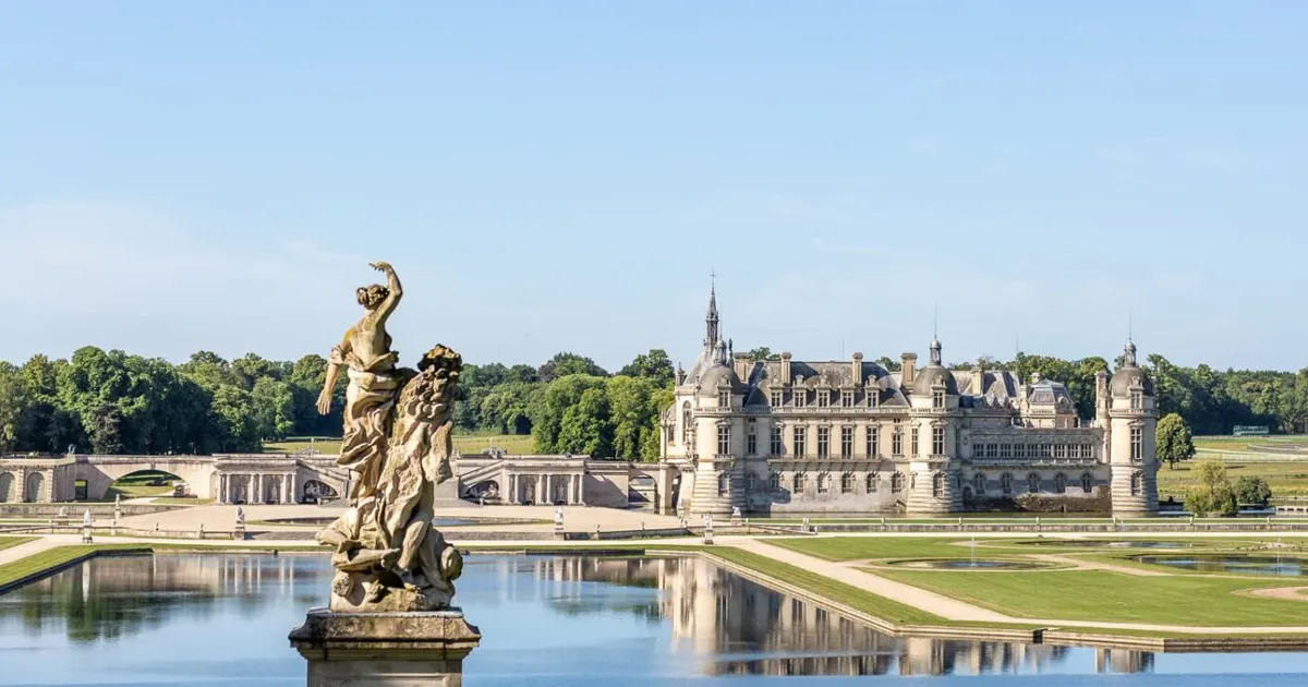 A sculpture of a woman in a fountain i the grounds of a large stone manor house in France.