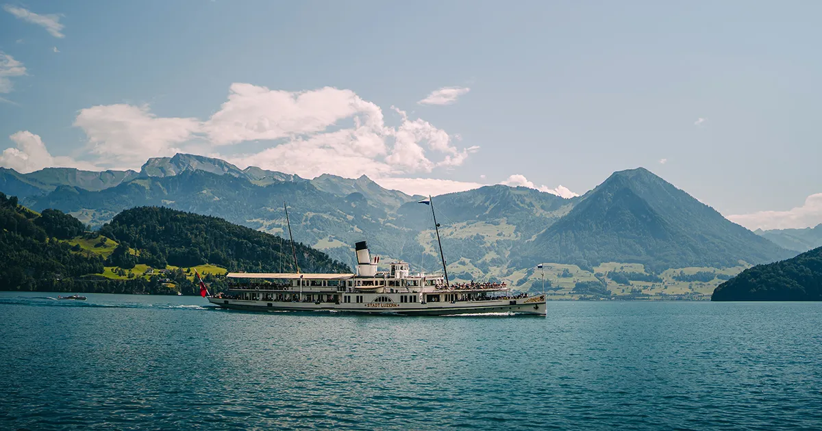 A steam boat cruises on a lake with a backdrop of mountains and farmland in the Swiss Alps.