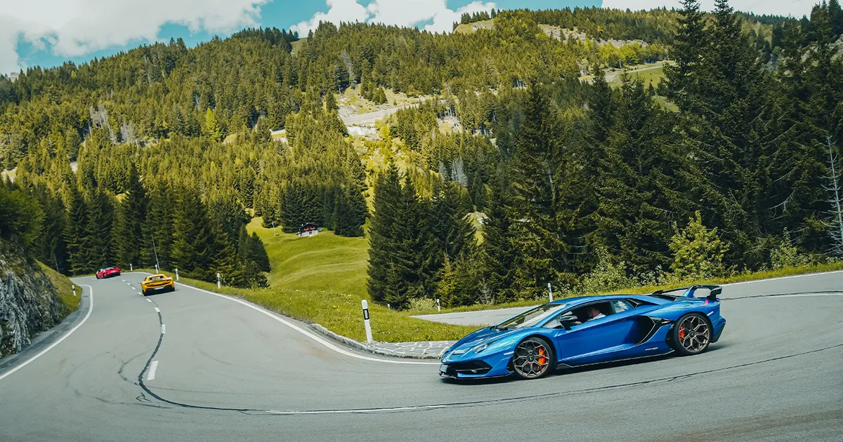 A blue Lamborghini Aventador rounds a tight corner on a green valley road in a pine forest 