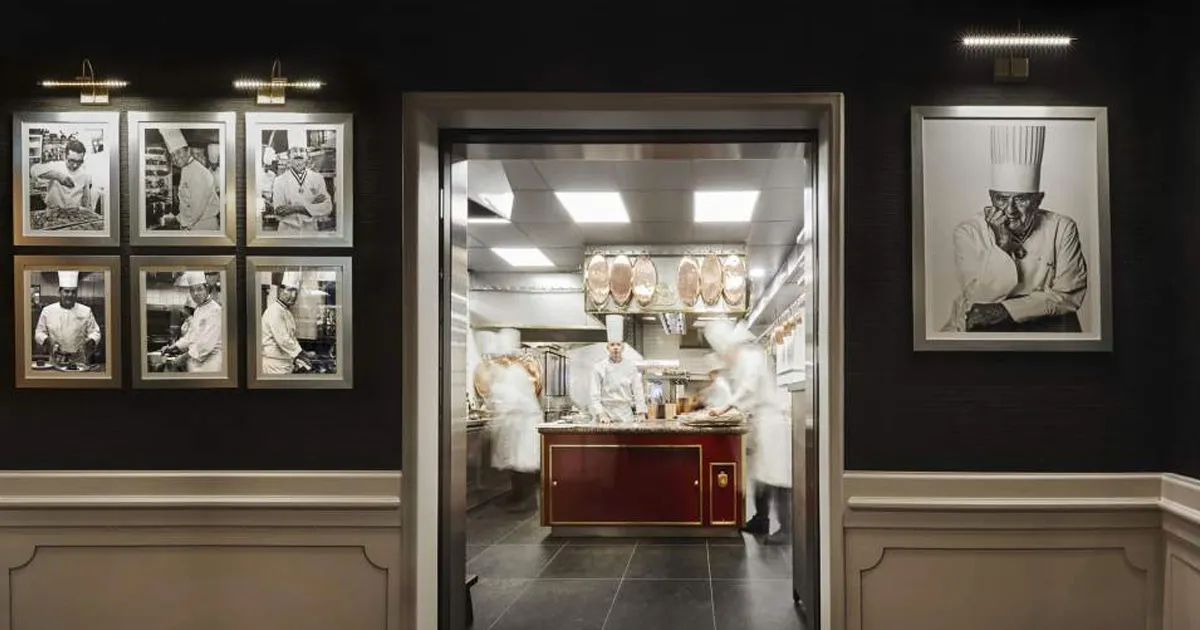 A hallway with photos and a doorway to a busy kitchen with chefs wearing whites and working busily.