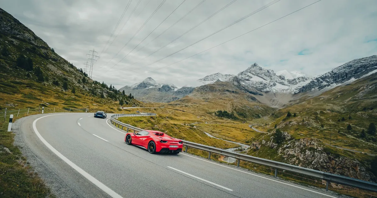 A red Ferrari 488 follows a blue Porsche 911 up an alpine pass