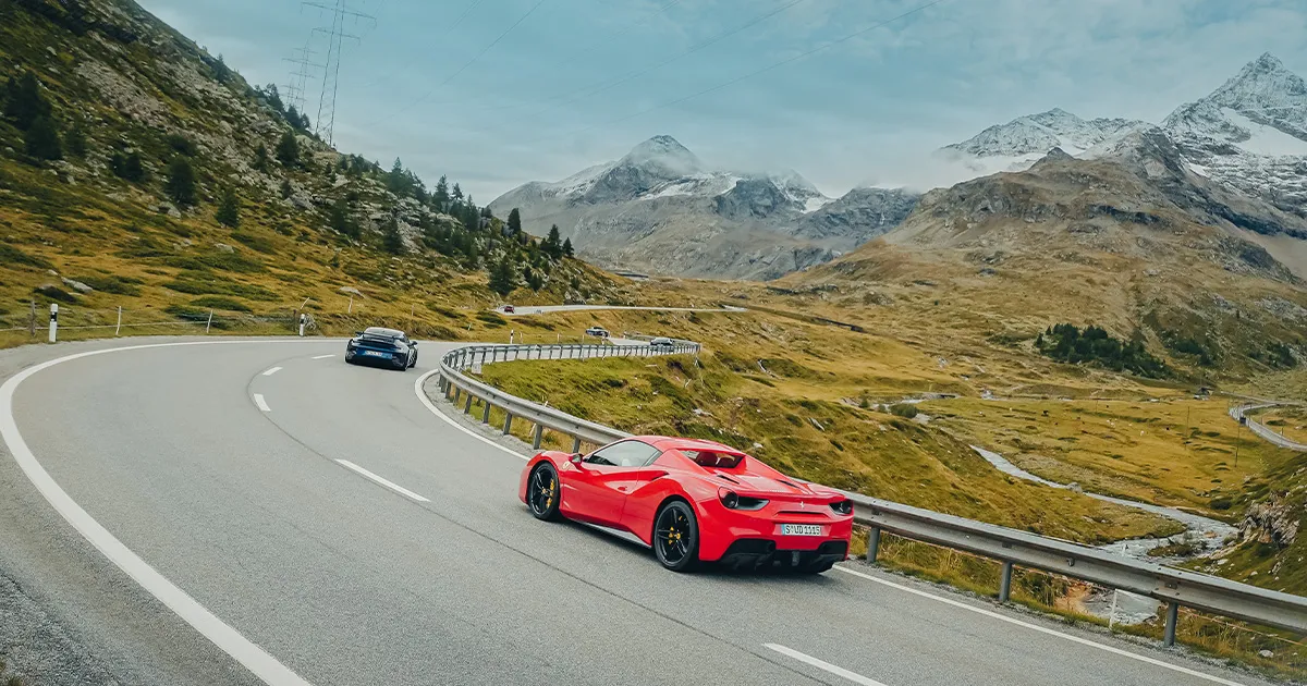 A red Ferrari follows a blue Porsche up an alpine road.