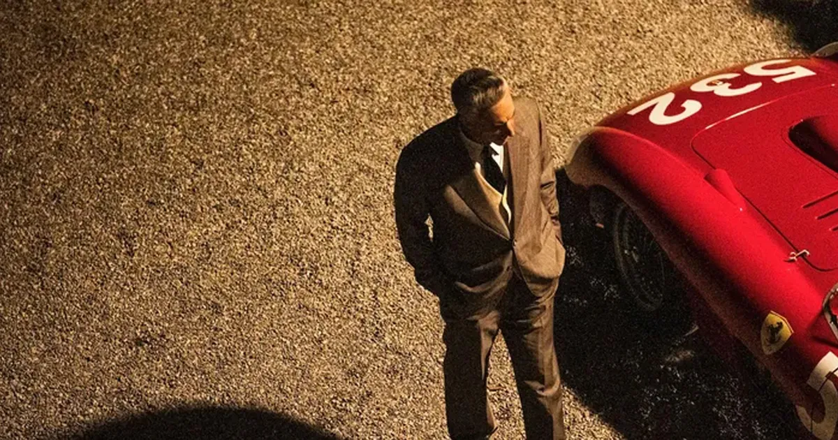 Adam Driver wearing a suit as Enzo Ferrari standing next to a racing car.