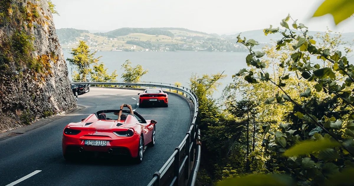 Two red Ferraris approach a corner on a road above a lake, as a passenger stretches above the windscreen to take a picture.