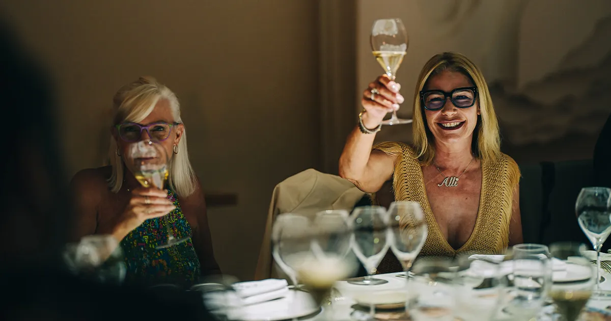 Two happy women wearing glasses share a toast at an elegant dinner table.