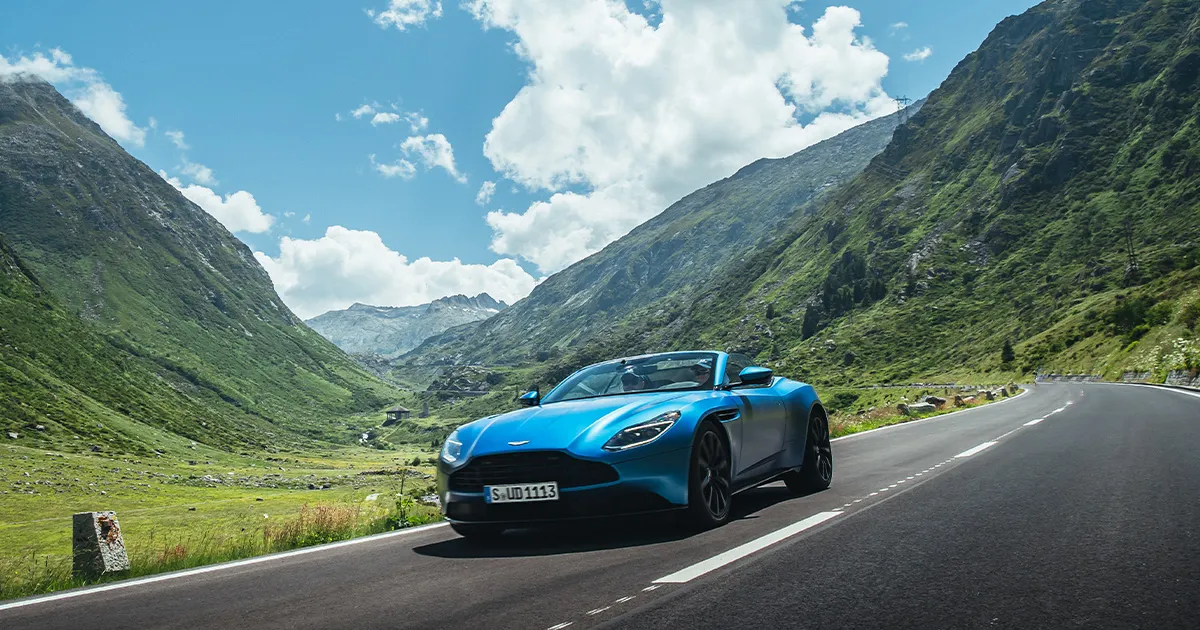 A bright blue Aston Martin convertible cruises along a high country road.