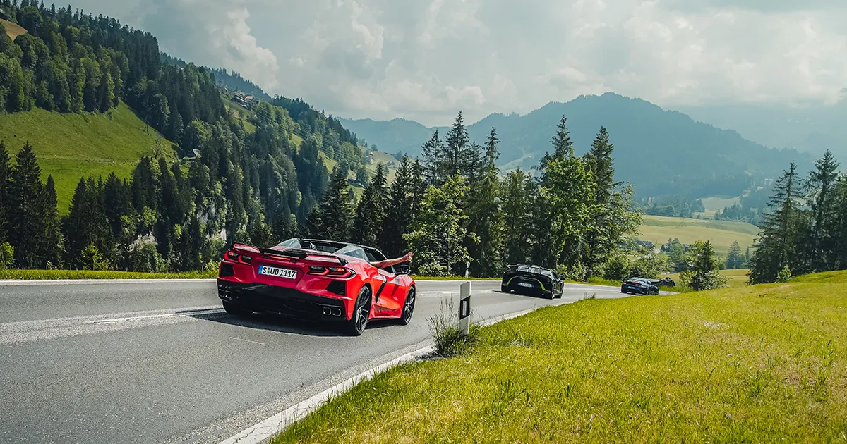 A red Chevrolet Corvette tails a brace of supercars along a green valley road.