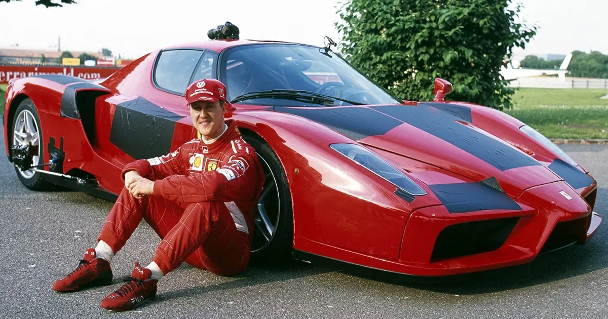 Michael Schumacher sitting in his racing uniform by a partially camouflaged red Ferrari Enzo supercar.