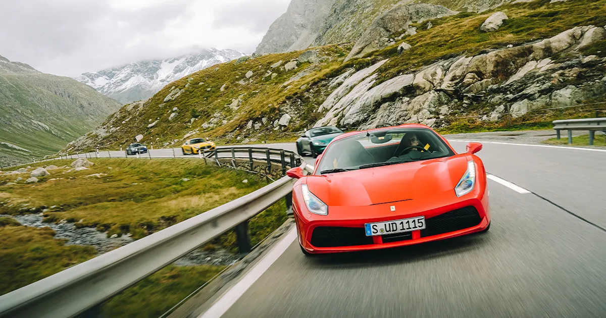 A red Ferrari 488 leads a green Aston Martin Vantage and two more supercars up an alpine pass.