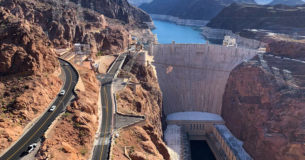 An enormous concrete dam in a red valley seen from a distance on a sunny day.