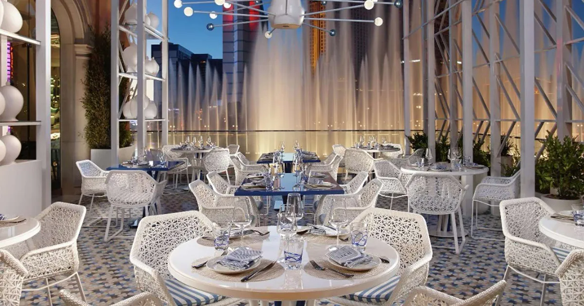 Luxurious white tables with striped trim sit on a patio overlooking a huge fountain.