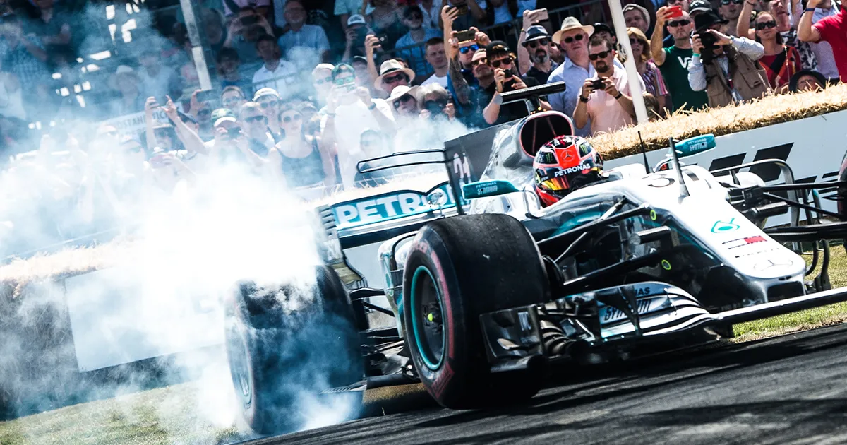 A driver performs a burnout with smoke rising from the rear tyres of a modern racing car with sponsor branding and green trim in front of an enthusiastic crowd at Goodwood Festival of Speed.