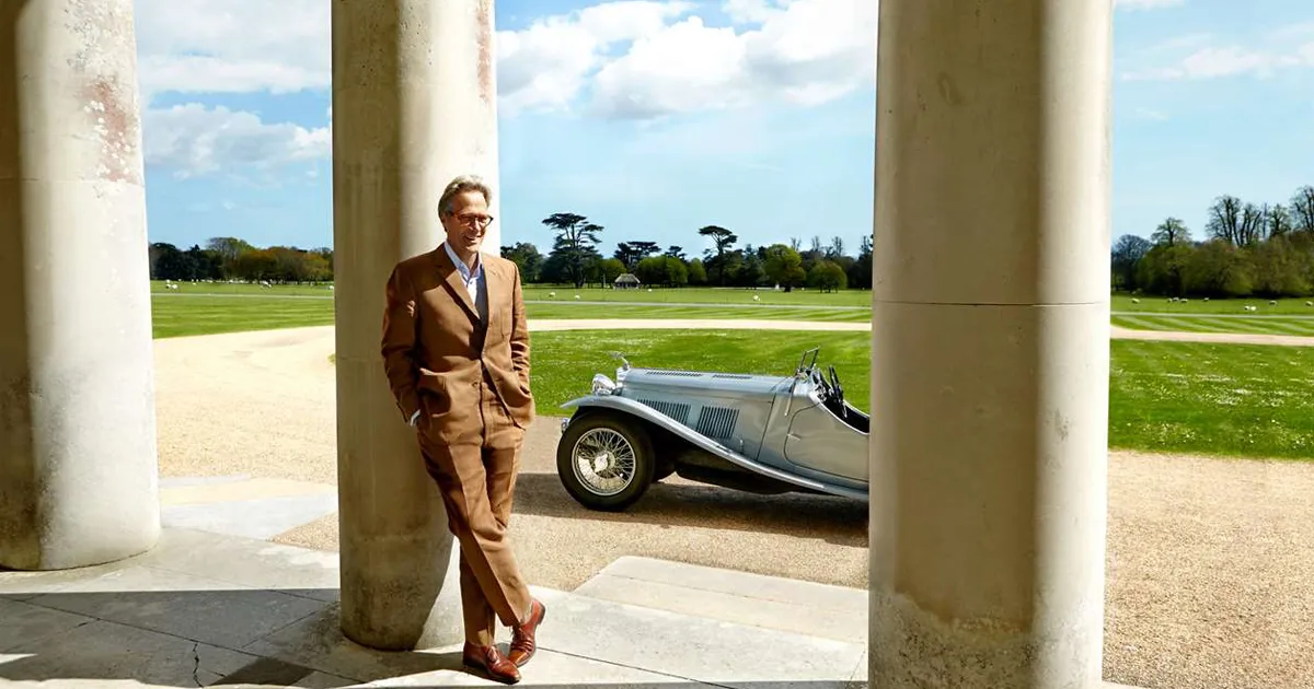A man with grey hair and glasses in a stylish brown suit stands relaxed in front of a silver classic car and sweeping green country grounds in Goodwood, England.