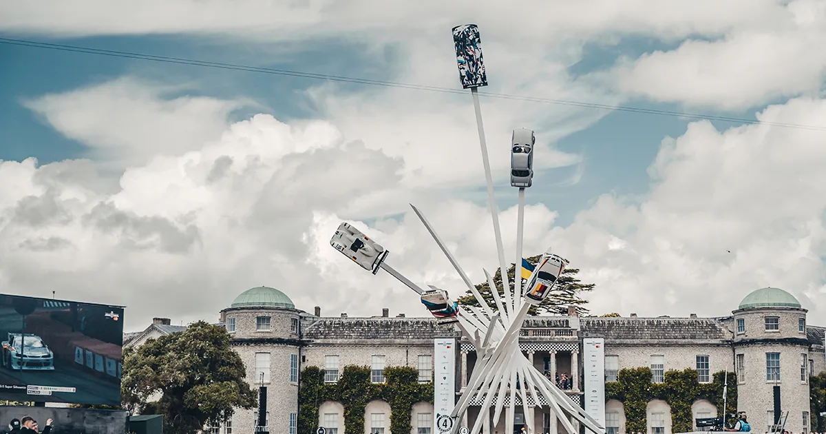 A huge white metal sculpture with racing cars on long girders in a stylish formation, in front of Goodwood house, England.