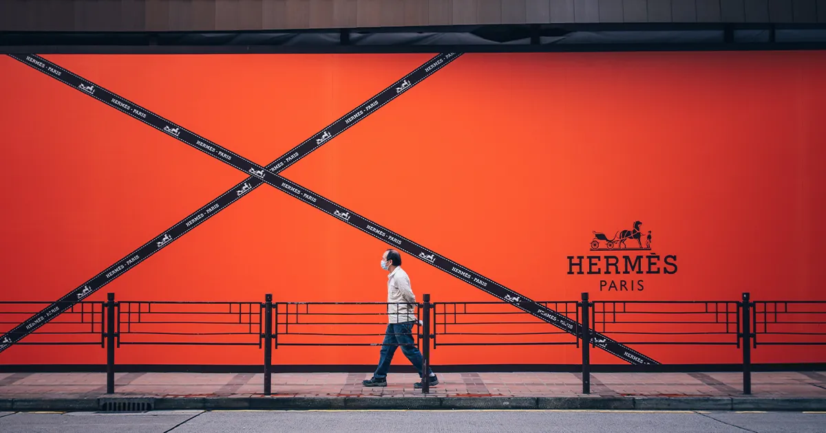 A man in a mask walks along a path with an orange background displaying the Hermes logo and a large ribbon.