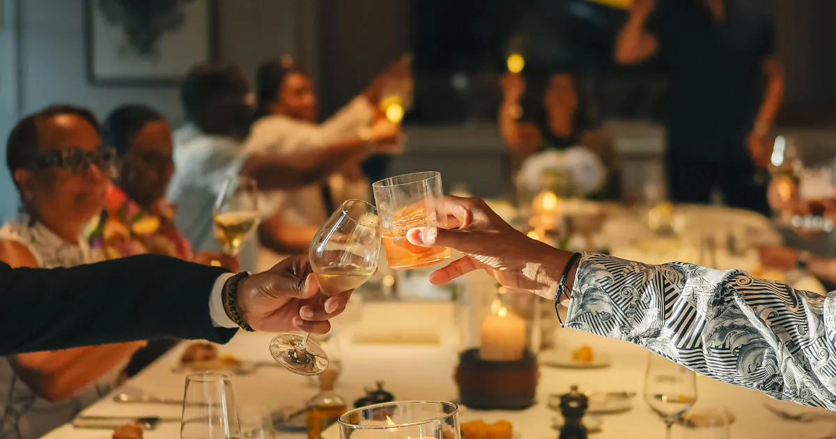 Group of people round a large dinner table cheers cocktail and wine glasses