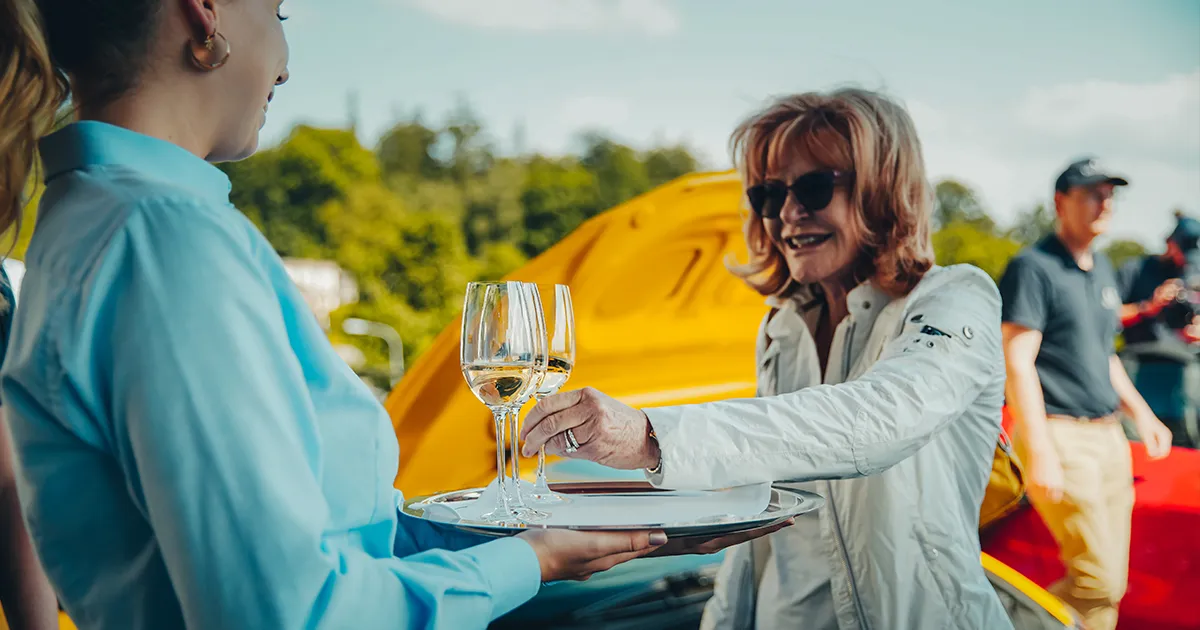 A lady in sunglasses takes a glass of white wine from a waitress’s tray.