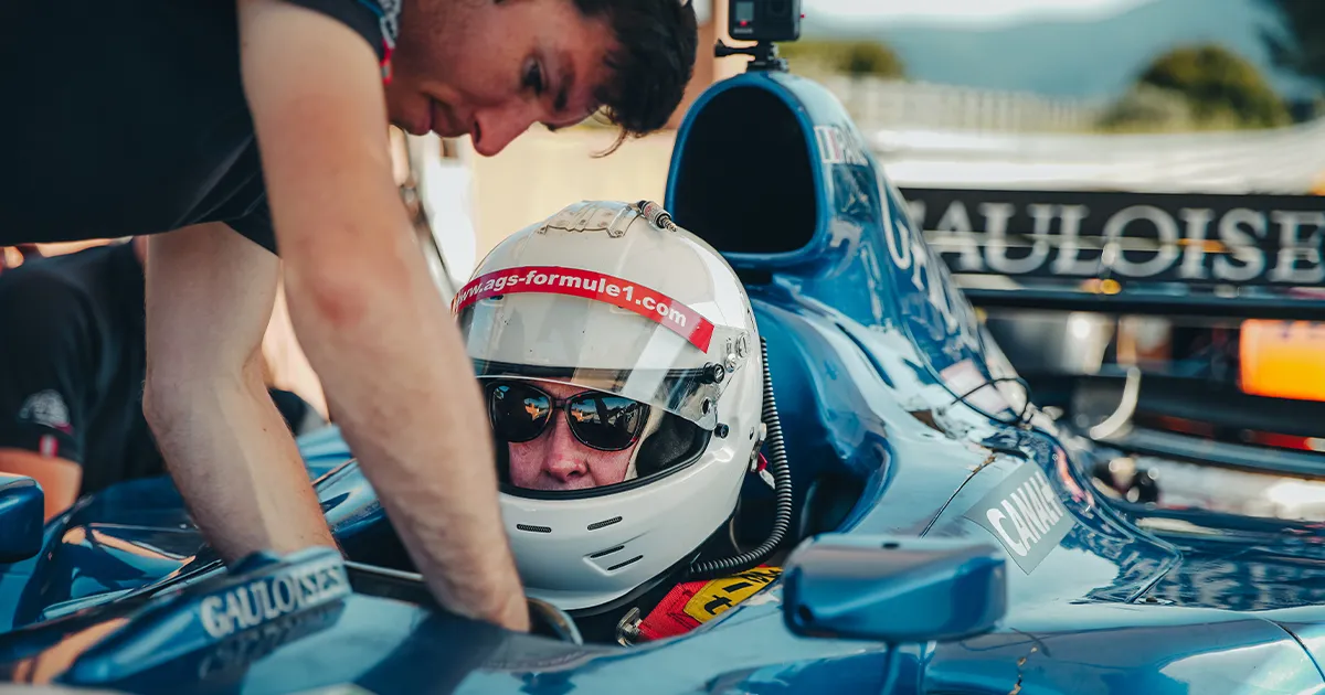 A person wearing sunglasses and a full face helmet is assisted as they sit in a blue Formula 1 car.