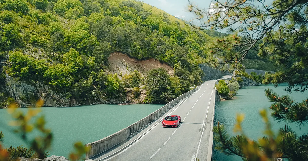 A red Lamborghini crosses a bridge over a large body of water.