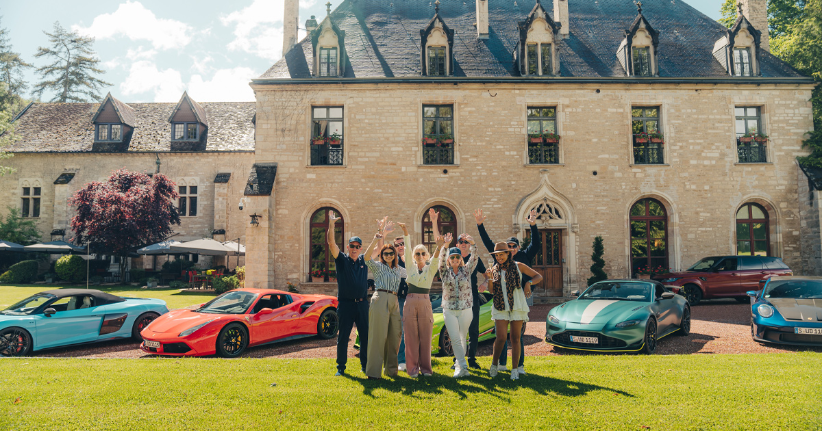 A small tour group posing with hands raised in front of supercars and a beautiful mansion