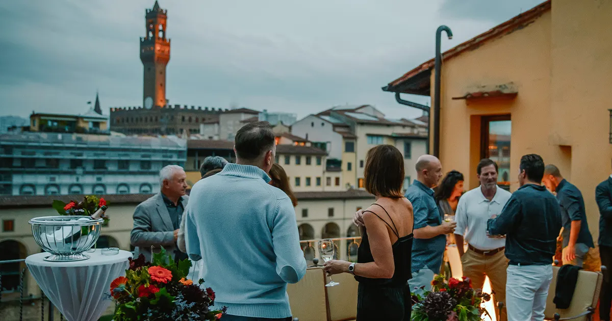 A group of smartly dressed party guests on a rooftop enjoying evening views across a historic city.