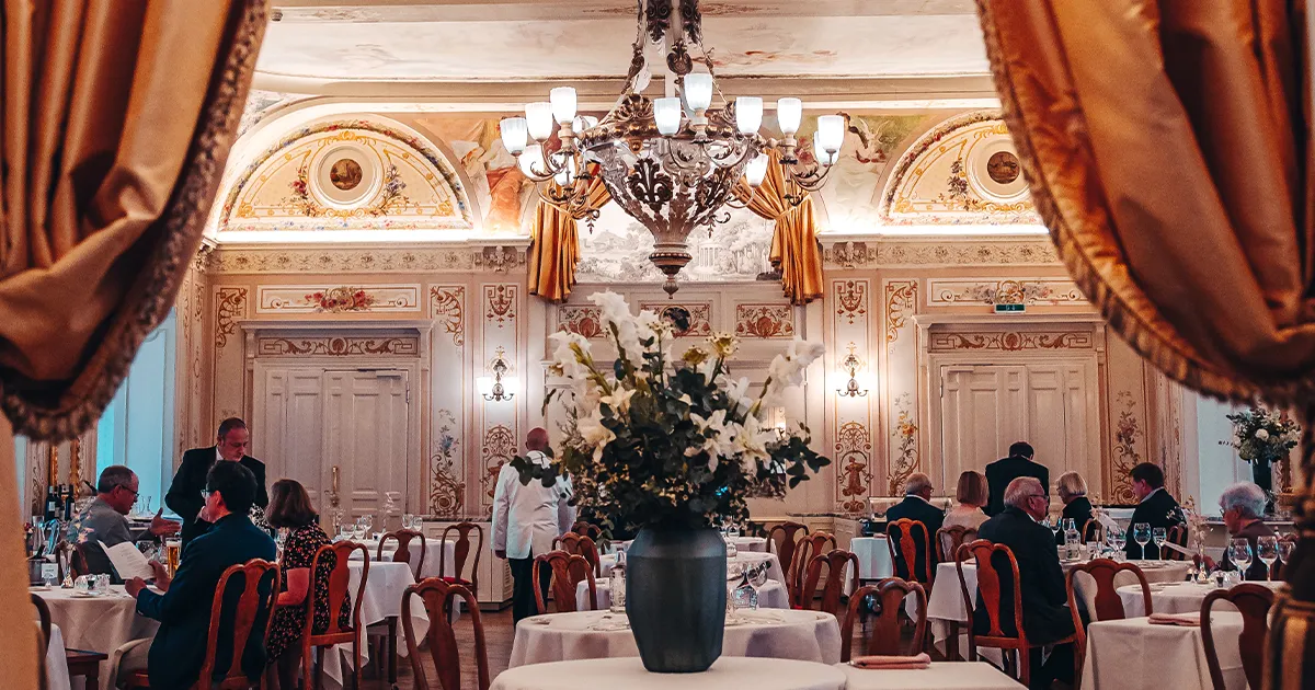 An ornate dining room with diners seated at round tables with patterned walls and large curtains.