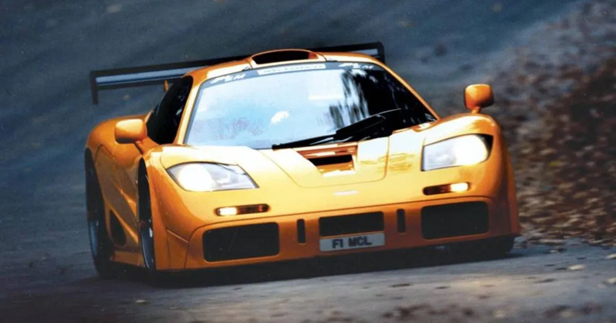 An orange McLaren F1 GTR with its headlights on as it is driven on a country road
