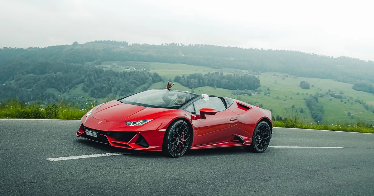 A man in the passenger seat of a red Lamborghin Huracan convertible has his hands raised in celebration.
