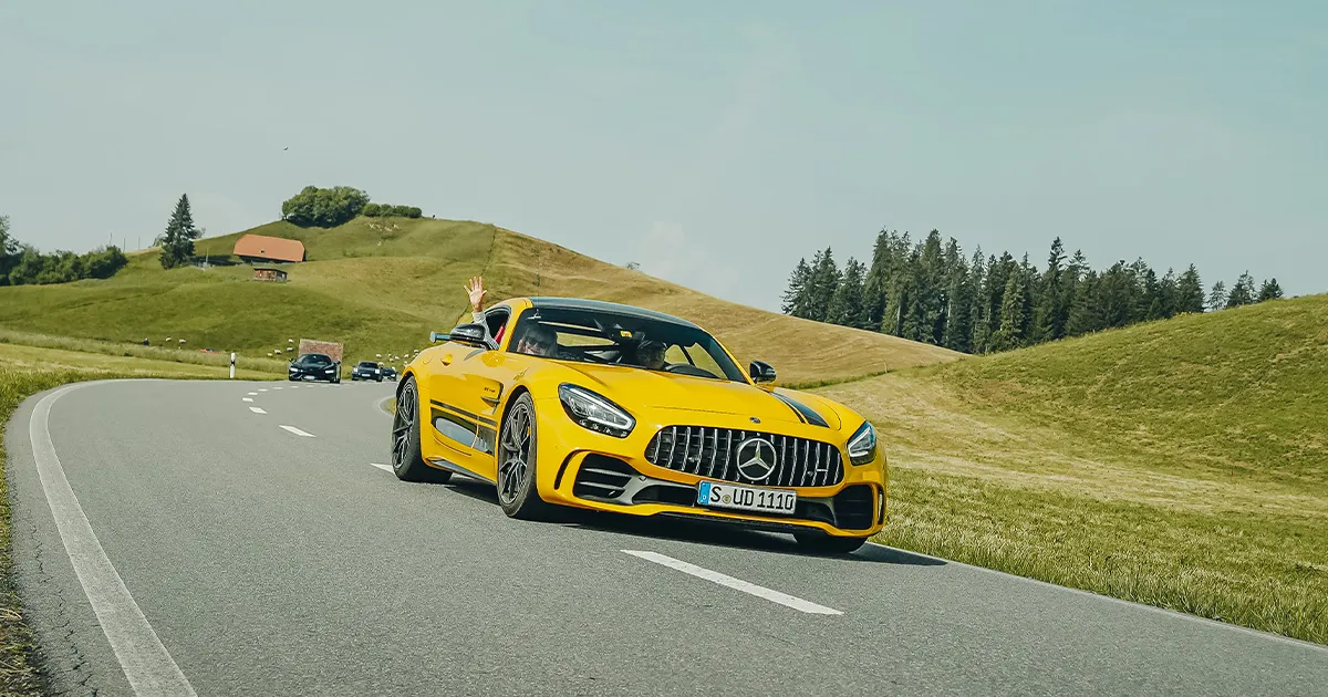 A yellow Mercedes-Benz AMG GTR driving on a country road with a female passenger waving to the camera.
