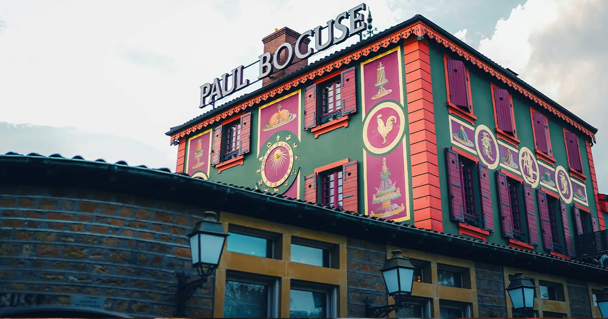 A decorative restaurant with wooden shutters, painted green and red with a large sign on its roof with the words Paul Bocuse