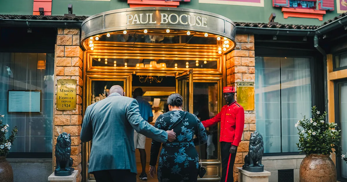 A couple are greeted by a bellhop who holds an ornate door open for them at Restaurant Paul Bocuse, Lyon.