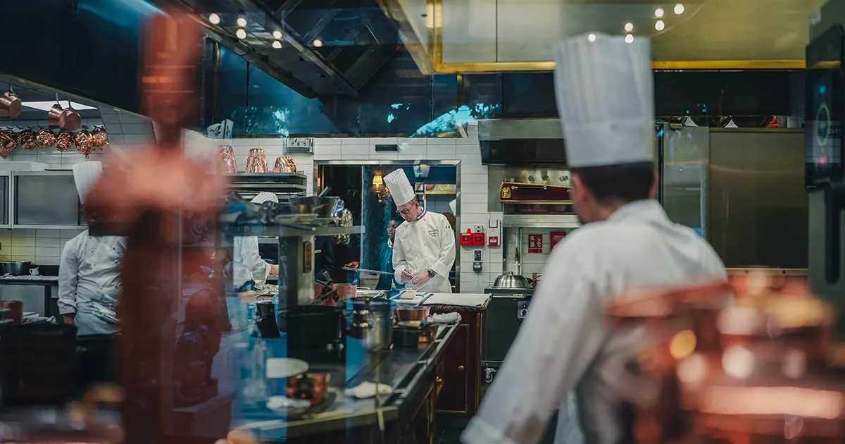 A chef wearing traditional whites inspecting a dish in the busy professional kitchen at Paul Bocuse.