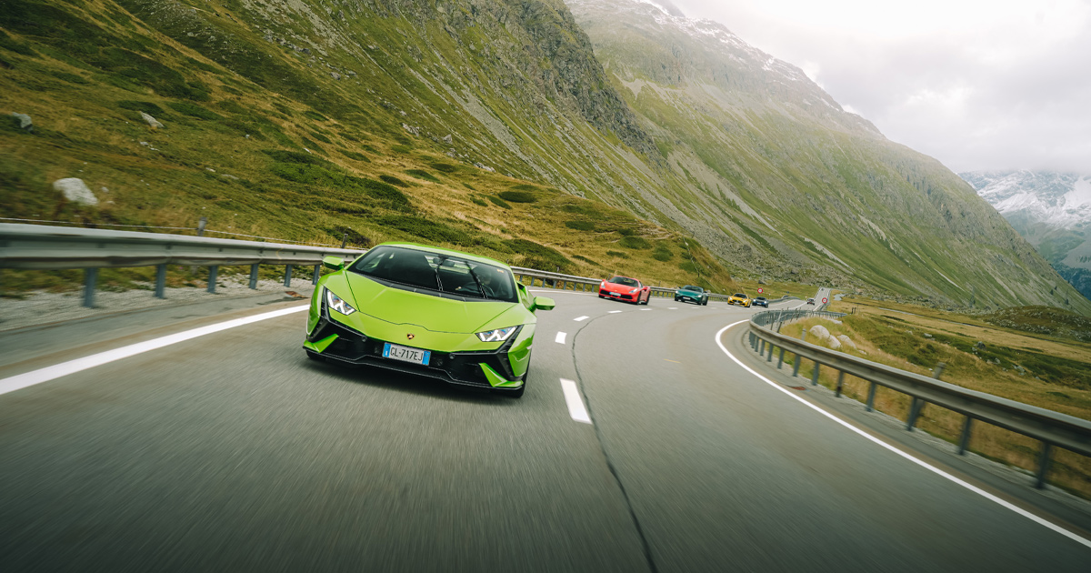 A green Lamborghini Huracan leading a red Porsche and other supercars round a bend in the Swiss Alps.