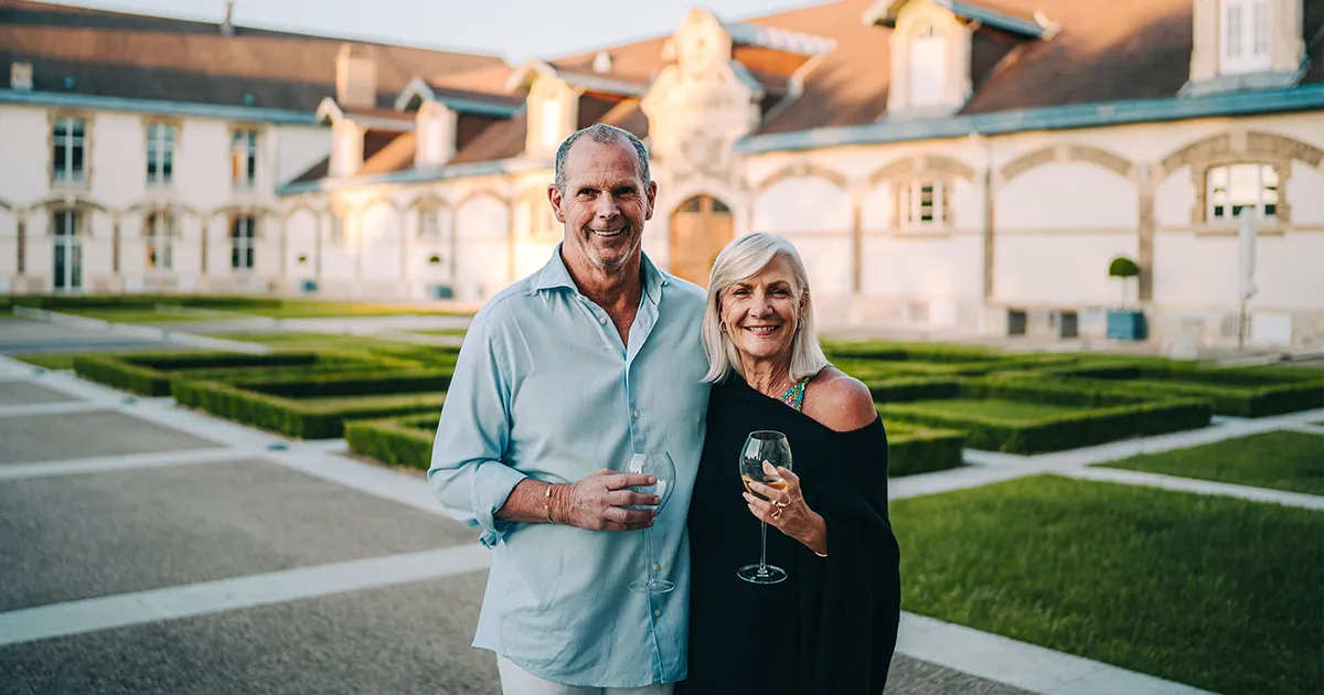 A happy man and woman sip wine in front of an opulent manor and garden.