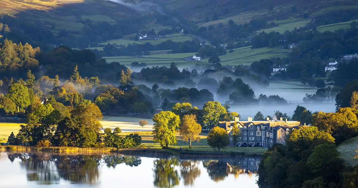 A wide shot of a stately home sitting atop a lake as morning mist rises in Northern England.
