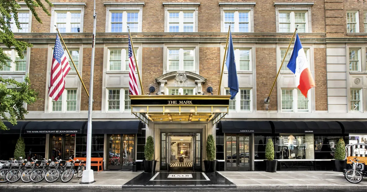 An exterior shot of the entranceway of a luxurious brick hotel with large flags above its doorway, The Mark, New York.