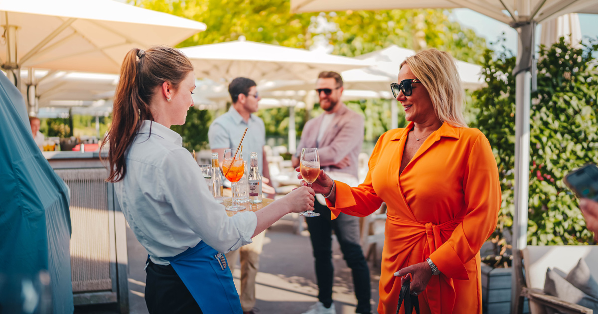 A waitress serves wine to a lady wearing a fashionable orange outfit with sunglasses 