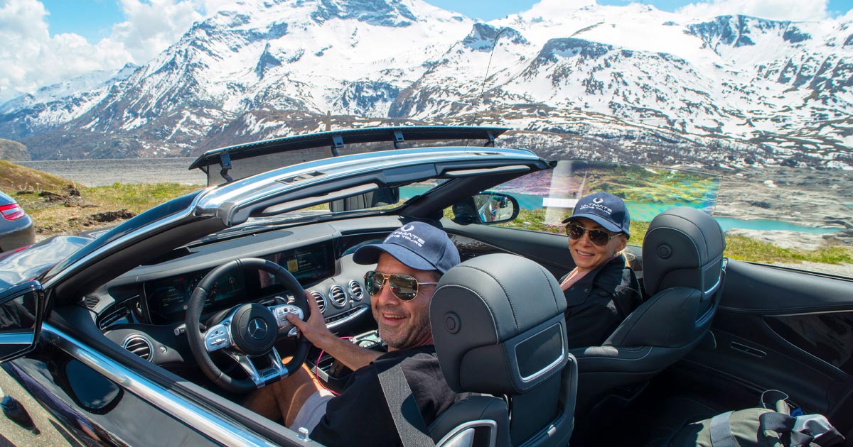 A happy couple decked out in branded hats enjoying a supercar in the Alps with Ultimate Driving Tours