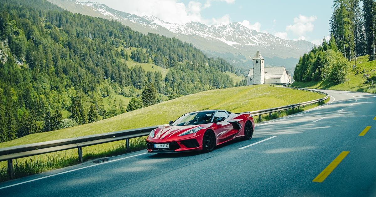 A red Chevrolet Corvette C8 rounds a gentle corner in a European alpine region