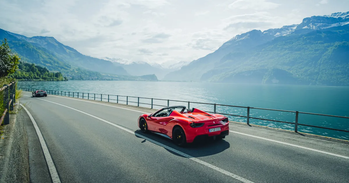 A red Ferrari 488 Spyder cruising on a highway next to a beautiful alpine lake